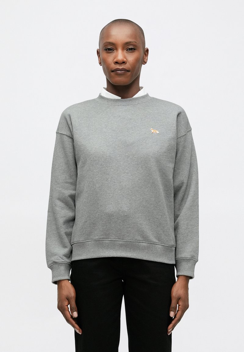 Sudadera gris de tejido suave, con cuello redondo, corte holgado y un logo de tigre bordado en el lado izquierdo del pecho. Pantalones negros.
