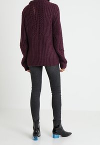 Maglione a maglia spessa di colore viola intenso con un alto colletto, caratterizzato da un motivo testurizzato e un dettaglio di taglio sulla schiena, abbinato a pantaloni grigi slim e stivaletti neri lucidi.