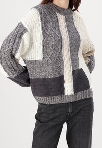 Suéter de punto con diseño de patchwork en gris y crema, que presenta patrones de trenzado y texturizados, puños acanalados y cuello redondo.