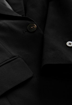Černý blazer vyrobený z hladké látky s klopovým límcem, se dvěma knoflíky – jedním tmavým a jedním světlým – a detailem kapsy s klopou.