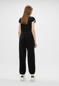 Czarny kombinezon z miękkiego materiału, z krótkimi rękawami i elastycznym paskiem w talii. Posiada zwężane nogawki oraz klasyczne mankiety.