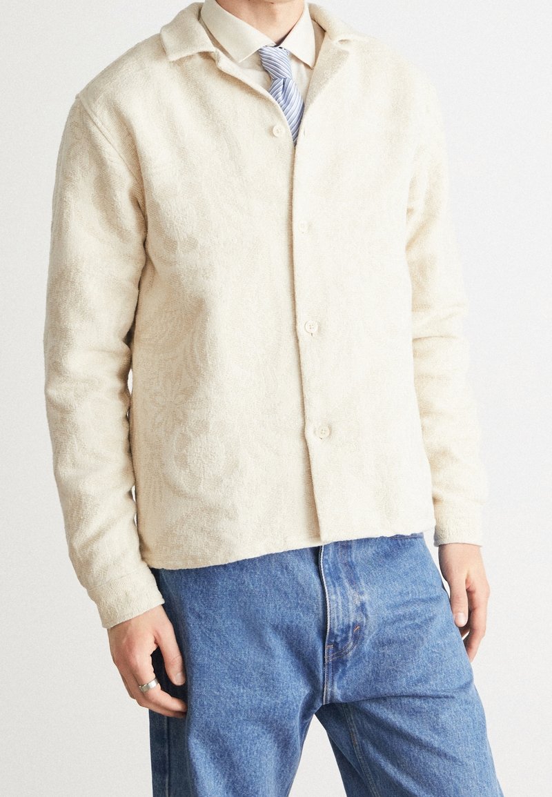 Beige strukturerad knappjacka med blommönster, kragdesign, bärs över en ljus skjorta, kombinerad med blå denimjeans.