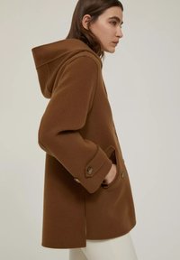 Cappotto di lana marrone con cappuccio, dotato di una fessura laterale, ampie tasche frontali e dettagli con bottoni. Texture liscia e forma su misura.