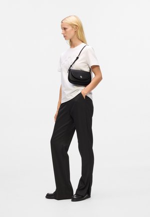 Black crossbody bag with a textured finish, featuring a curved shape, zip closure, and a subtle logo accent. Worn with a white shirt and black pants.