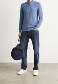 Hellblauer Reißverschluss-Pullover, dunkle Jeans und weiße Sneakers. Hält eine runde marineblaue Tasche mit einem goldenen Hardware-Detail. Gesamte glatte Textur.
