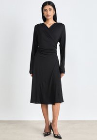 Black wrap dress with long sleeves, smooth texture, knee-length hem, and fitted waist, paired with black heels featuring gold accents.