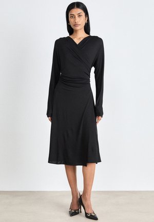 Black wrap dress with long sleeves, smooth texture, knee-length hem, and fitted waist, paired with black heels featuring gold accents.