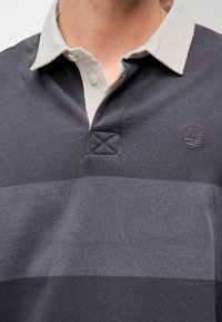 Gros plan sur une personne portant un maillot de rugby gris avec un col beige clair et un logo d'arbre sur la poitrine.