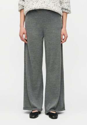 Persona che indossa pantaloni in maglia grigi a gamba larga, scarpe piatte nere e un maglione chiaro maculato, in piedi contro uno sfondo semplice.