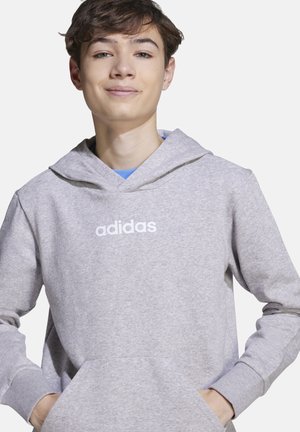 Adolescent portant un sweat à capuche Adidas gris clair, les mains dans la poche avant, souriant légèrement, sur un fond clair uni.