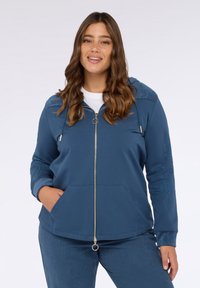 Blaue Zip-Hoodie mit gefütterter Kapuze, vorderen Taschen, gesticktem Muster an den Ärmeln und einem goldenen Reißverschlussanhänger. Weicher Stoff.