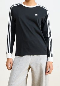 Černé tričko s dlouhým rukávem, bílé proužky na rukávech, kulatý bílý límec a malá vyšívaná adidas logo na hrudi.