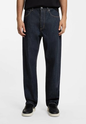 Mann trägt dunkelblaue Jeans mit geradem Bein und schwarze Sneaker mit weißen Sohlen, steht vor einem einfachen Hintergrund.