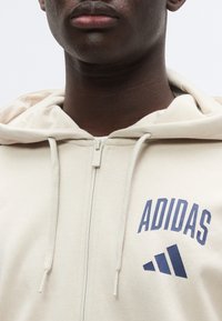 Μπεζ φούτερ με φερμουάρ, κατασκευασμένο από απαλό ύφασμα, με μεγάλο μπλε λογότυπο "ADIDAS" και διακόσμηση με τρεις ρίγες. Περιλαμβάνει κορδόνια στη κουκούλα.