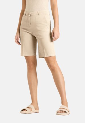 Vrouw die beige knielange shorts en lichtbeige gevlochten sandalen draagt, staand tegen een effen witte achtergrond.