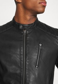 Svart läderjacka med hög krage, vadderade axeldetaljer och en dragkedjefrontficka med metalliska accenter.