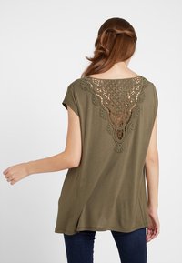 Blusa verde olivo de manga corta. Presenta un detalle de ganchillo en la espalda, creando un patrón decorativo de recortes. Textura de tela suave.