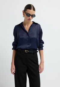 Camicia di seta blu marina con colletto pieghevole e bottoni davanti, abbinata a pantaloni neri e una cintura nera strutturata con un accento dorato.