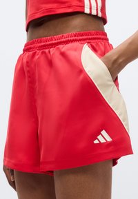 Κόκκινα αθλητικά σορτς με ελαστική μέση, με κρεμ πλαϊνή λωρίδα και λευκές λεπτομέρειες με το λογότυπο Adidas. Λείο, ελαφρύ ύφασμα.