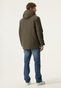 Olijfgroen pufferjack met een capuchon, met een soepele textuur en verstelbare trekkoorden, gecombineerd met blauwe jeans en lichte schoenen.