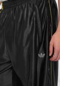 Melnas sporta bikses ar spīdīgu tekstūru, ar dzeltenu sānu strīpu, snaps pogām un mazu pelēku adidas logotipu uz kājas.