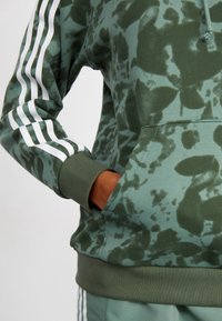 Green hoodie made of soft fabric featuring a splatter pattern, kangaroo pocket, and white striped accents on sleeves.