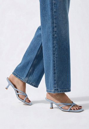 Pies con sandalias de tacón de tiras azul claro combinadas con vaqueros azules de corte recto sobre un fondo liso.