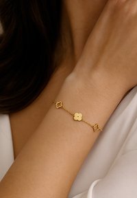 Auksinis apyrankė su gėlių formos jungtimis, alternatyviais tvirtais ir atvirais dizainais, sujungiami švelnia grandinėle, ilsisi ant riešo.