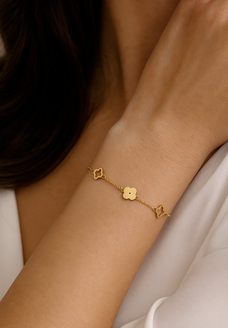 Auksinis apyrankė su gėlių formos jungtimis, alternatyviais tvirtais ir atvirais dizainais, sujungiami švelnia grandinėle, ilsisi ant riešo.