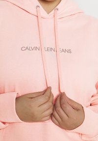Sudadera de algodón rosa con cordón, que presenta un logo "CALVIN KLEIN JEANS" en gris claro. Se aprecian detalles de puños y dobladillo acanalados.