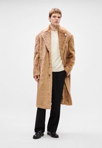 Brūns mākslīgā kažokādas mētelis ar plašiem atloka, pogu aizdari un kabatām. Valkāts virs krēmkrāsas ribota džempera un melnām biksēm.