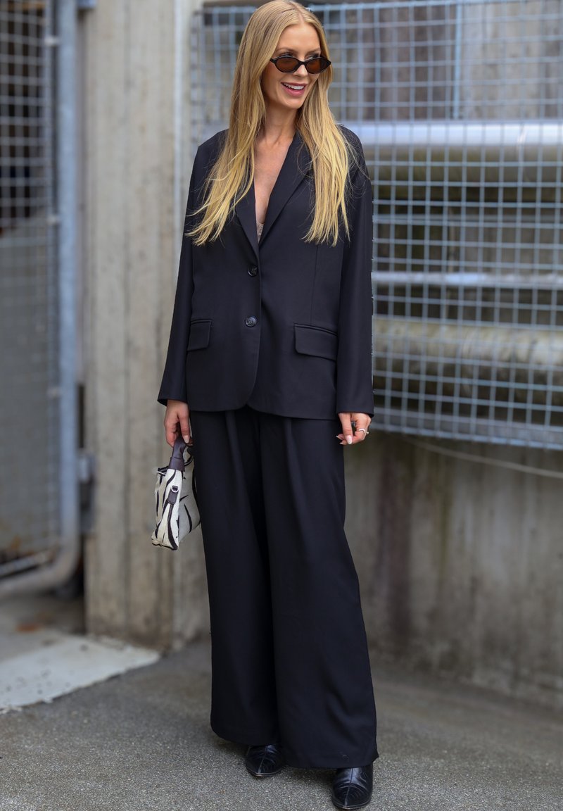 Femme aux longs cheveux blonds portant un costume noir oversize et des lunettes de soleil, tenant un petit sac à main à motifs, debout près d'une clôture métallique en extérieur.