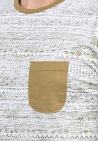 Valkoinen t-paita, jossa on teksturoitu, geometrinen kuvio kullan ja beige sävyissä, ja vasemmassa rintakehässä on kiinteä beige tasku.
