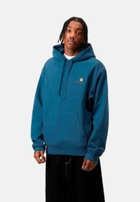 Sudadera azul de algodón con un bolsillo frontal, capucha con cordón y pequeño detalle de logo en el pecho. El modelo lleva pantalones oscuros.