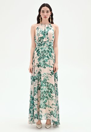 Donna che indossa un abito maxi beige senza maniche con stampa floreale verde, abbinato a scarpe bianche con punta e orecchini dorati vistosi.