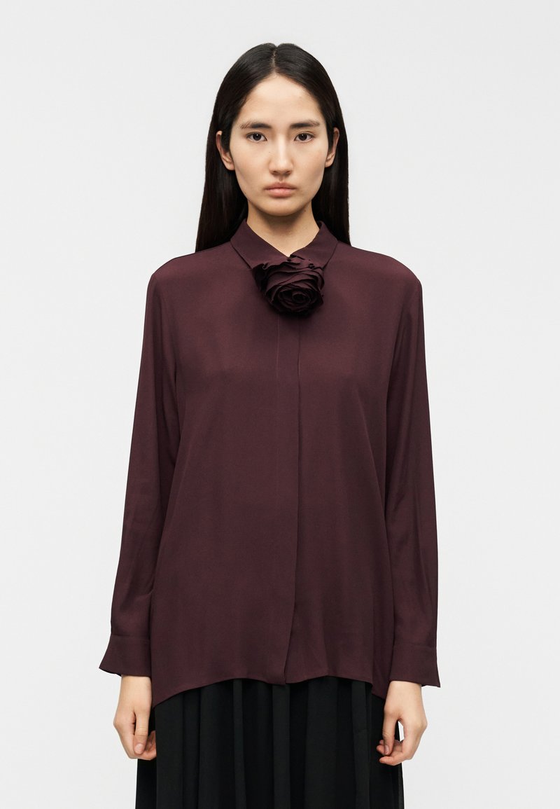 Μπορντό μπλούζα με μακριά μανίκια από υφασμάτινη ύφανση, με σκαλιστή λεπτομέρεια τριαντάφυλλου στο γιακά. Ίσια γραμμή στο τελείωμα και κουμπιά στο εμπρός μέρος.