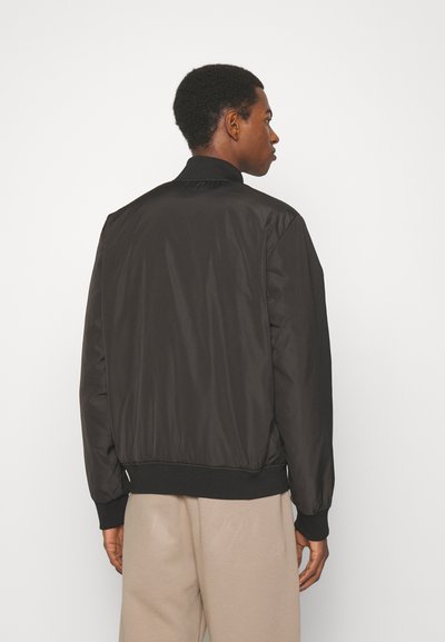 Veste bomber noire en matériau lisse et léger, avec un col et des poignets côtelés. Présente une taille ajustée et un design minimaliste.