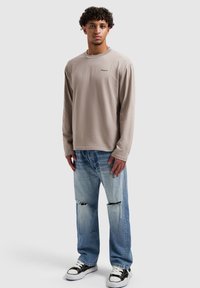 Beige shirt met lange mouwen en geborduurd logo, gecombineerd met lichtblauwe gescheurde spijkerbroek en zwart-witte sneakers. Eenvoudig, casual ontwerp.