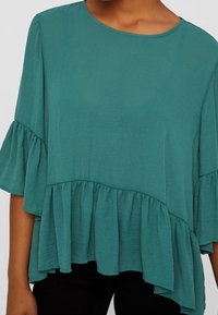 Blusa leggera color turchese con scollatura rotonda, maniche corte a balze e orlo a balze. Tessuto morbido e vestibilità ampia.