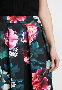 Květinová plisovaná sukně se černým základem a jasně růžovými a bílými růžemi, zelenými listy a hladkou texturou. Viditelný úzký pas.