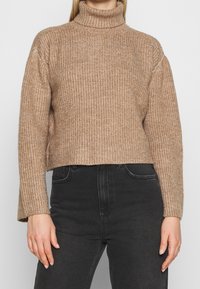 Beige tröja med ribbad textur, hög polokrage och kort design. Matchad med svarta jeans, som har en avslappnad passform och sidofickor.