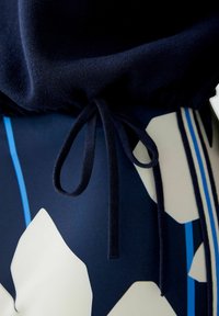 Marineblauer Strickpullover mit gerafftem Saum und Kordelzug. Kombiniert mit einem gemusterten Stoff in Creme und blauen geometrischen Formen.