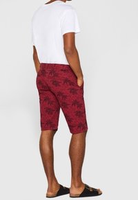 Shorts rouges à motifs avec des designs de palmiers, en tissu léger, associés à un t-shirt blanc et des sandales noires.