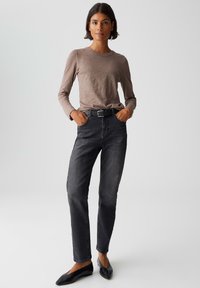 Una donna sta in piedi con le mani in tasca, indossando una maglietta a maniche lunghe color taupe, una cintura nera, jeans neri sbiaditi e ballerine nere a punta.