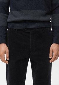 Maglione a strisce blu scuro con texture a coste, abbinato a pantaloni in corduroy neri con bottone e passanti per cintura. Mani visibili ai lati.