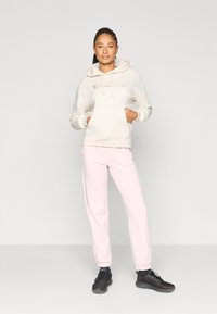 Ανοιχτό μπεζ φούτερ με κορδόνια και μπροστινή τσέπη, συνδυασμένο με ροζ παντελόνι φόρμας και μαύρα αθλητικά παπούτσια.