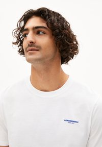 Witte katoenen t-shirt met korte mouwen, met blauwe geborduurde tekst "ARMEDANGELS SINCE 2007" op de linker borst. Ronde halslijn.