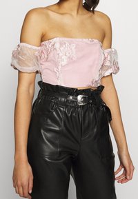 Crop top rose avec des manches transparentes dénudant les épaules, broderie florale, associé à un pantalon en cuir noir taille haute avec une taille ceinturée.