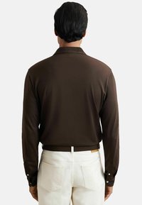 Brūna garām piedurknēm krekls ar pogātām aprocēm, valkāts ar baltām biksēm. Krekls ir ar gludu tekstūru un klasisku apkakles dizainu.