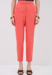 Coral tailored trousers with a slim fit, featuring side pockets and sharp creases down the front. Paired with brown shoes.